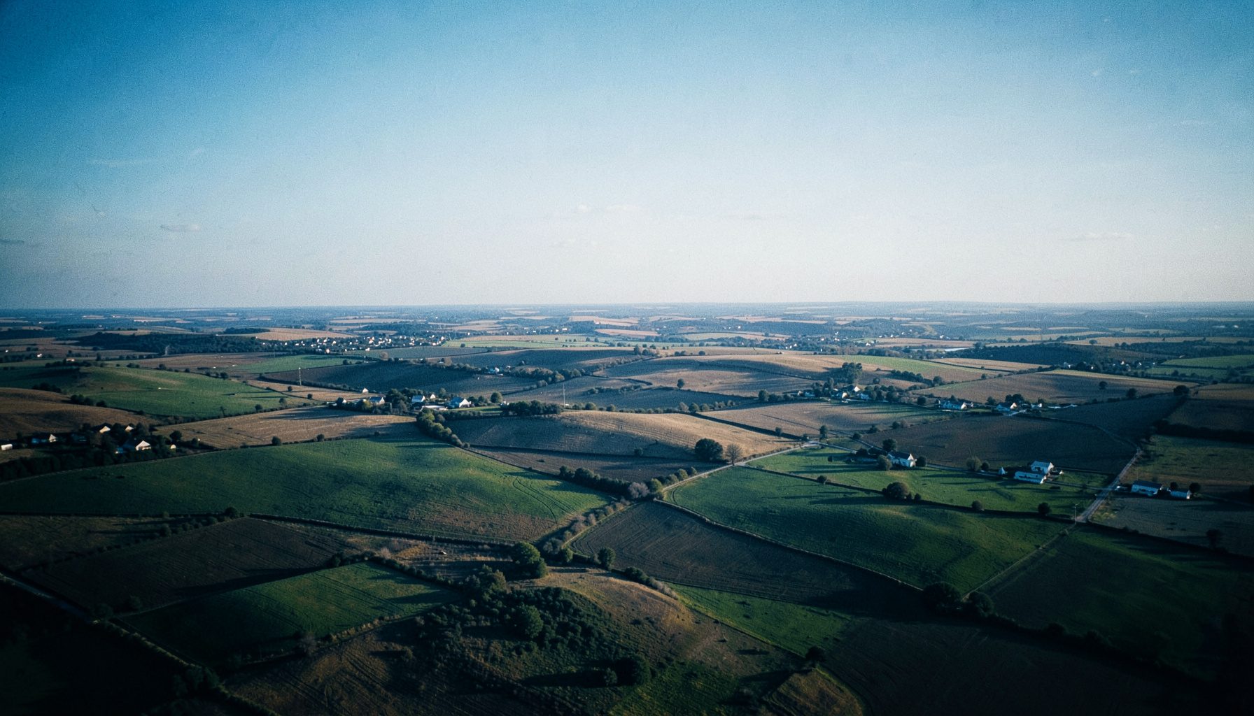 Wide aerial view of rolling Kansas landscape with patchwork fields and small towns scattered across the horizon under soft afternoon light